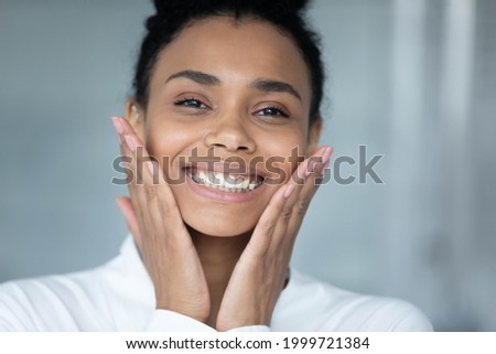 Head shot of happy joyful Black girl touching face in bathroom, admiring cosmetic effect of moisturizing cream, organic oil, cleaning lotion, looking at camera, smiling. Skincare, dental care concept