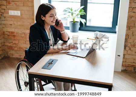 Young disabled business woman in wheelchair working at office desk and with laptop, talking on mobile phone accessibility and independence concept Royalty-Free Stock Photo #1999748849