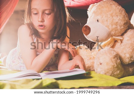Young beautiful girl reading a large book. Nearby  on the floor sits a Teddy bear. Concept of relaxation and friendship. Horizontal image.