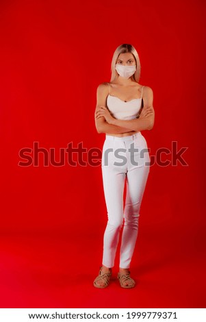 Serious standing blonde woman in face mask. Copy space photo of slim blonde in white medical mask and white outfit on matte red background. Blonde hair girl with outstretched arm holding something. 