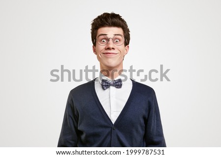 Cheerful young man in classic elegant suit with bow tie and nerdy glasses making funny grimace while smiling and looking at camera against white background