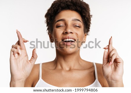 Young black woman in tank top holding fingers crossed for good luck isolated over white background