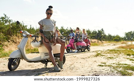 Beautiful young african american woman resting on retro scooter outdoors in warm sunny day with company of friends on background, widescreen. Leisure, fun and entertainment concept