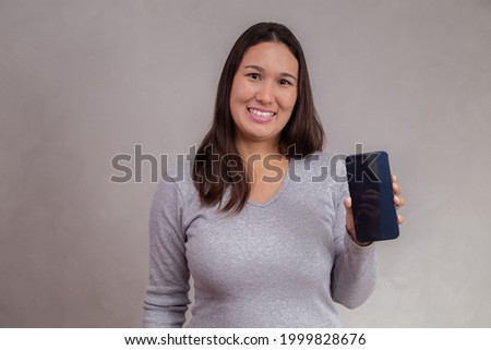 Smiling Asian woman is pointing on smartphone while standing over gray background.