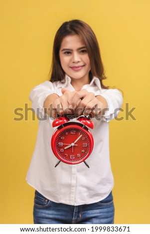 Smiling young Asian woman holding red alarm clock isolated on yellow background. Selective focus