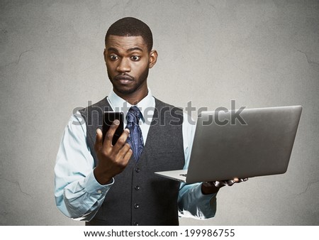 Closeup portrait shocked, surprised business man reading bad news on smart, mobile, cell phone holding laptop computer isolated black grey background. Human face expression emotion corporate executive