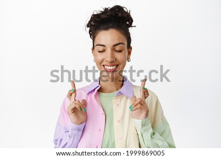 Smiling hopeful brunette girl makes wish, cross fingers and close eyes, anticipating results, waiting for dream come true, praying, standing over white background