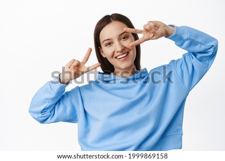 People and lifestyle. Young happy female in blue hoodie, showing peace v-signs, disco gesture near eyes, dancing and smiling joyful, standing over white background