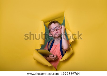 Smart child in glasses holding laptop computer tablet and breaking through yellow paper wall. Back to school. Student with backpack. Nerd kid portrait. Programming IT and developer lessons. Royalty-Free Stock Photo #1999869764