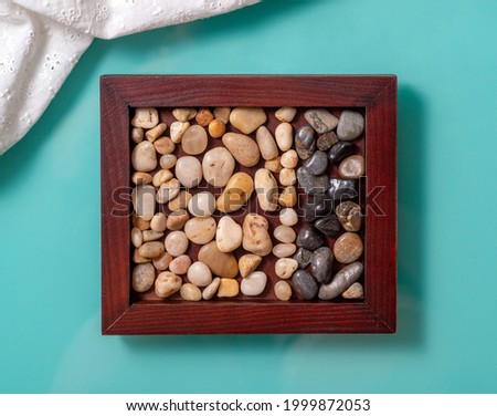 A wooden frame with sea pebbles on a turquoise background. The stones are lined with two stripes of light and dark, left in the corner a fragment of white fabric. View from above.