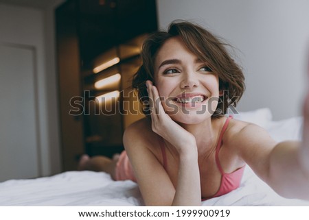 Close up young minded woman 20s wearing pajamas pink tank top shirt lying in bed do selfie shot on mobile cell phone rest relax indoors at home looking aside Good mood night morning bedtime concept.