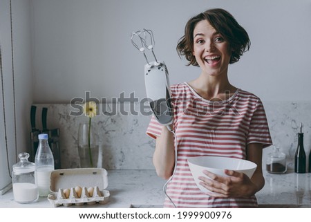 Young fun excited brunette housewife woman 20s in casual clothes striped t-shirt using mixer whips yolks beats eggs cooking food in light kitchen at home alone. Healthy diet bakery lifestyle concept Royalty-Free Stock Photo #1999900976