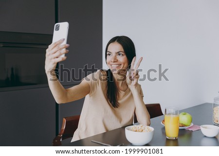Young housewife woman in casual clothes beige t-shirt eat breakfast talk by video call mobile cell phone show victory gesture cook food in light kitchen at home alone Healthy diet lifestyle concept