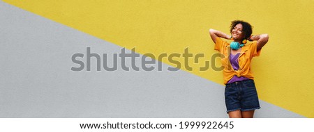 Positive woman with vitiligo wearing in stylish clothes and headphones stand near city wall painted in Ultimate Gray and Illuminating Yellow . Colors of 2021 year