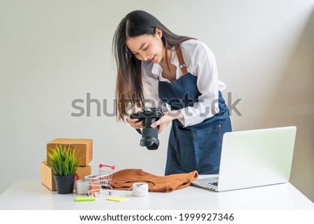 Asian woman business owner holding a camera to take pictures of products to sell online a laptop and parcel box at home.