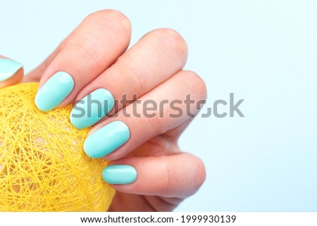 Female hand with beautiful manicure - turquoise, mint blue nails with a ball wrapped in yellow threads on blue background with copy space. Nail care concept