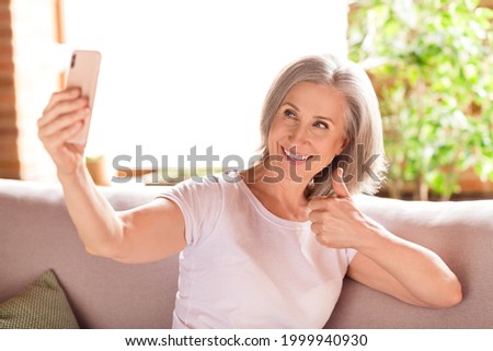 Portrait of elderly retired trendy cheery grey-haired woman sitting on sofa taking selfie showing thumbup at home house indoors
