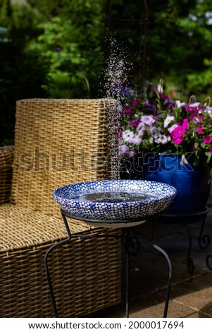 Beautiful patio with a rattan lounge chair, flower pots packed with flowers and a blue and white large pasta bowl filled with water and a solar pump which sprays up water to the delight of the birds.