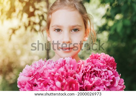 portrait of a happy cute little caucasian seven year old kid girl, holds in hands a bouquet of pink peony flowers in full bloom on the green background of nature. flare