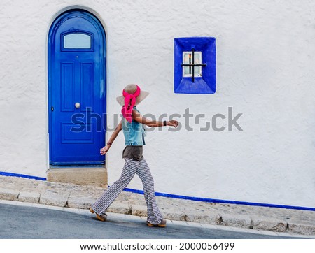 a caucasian woman walks quickly down a street in front of a white facade with blue doors and windows. she stands out against the white background. summer concept and tourism in Sitges, Spain.