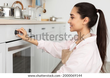 Household Concept. Closeup side view portrait of millennial cheerful woman in apron using stove in modern kitchen at her new apartment, holding and turning knob, controlling temperature of the burner Royalty-Free Stock Photo #2000069144