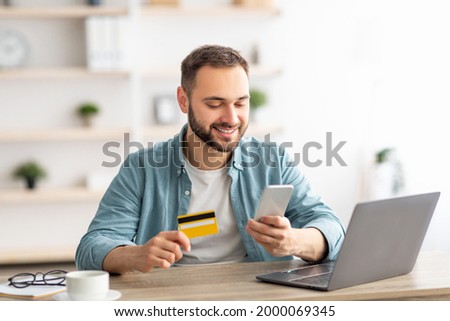 Happy Caucasian man buying things online, using smartphone, laptop and credit card, enjoying shopping in internet. Positive young guy purchasing goods on web, making remote payment Royalty-Free Stock Photo #2000069345