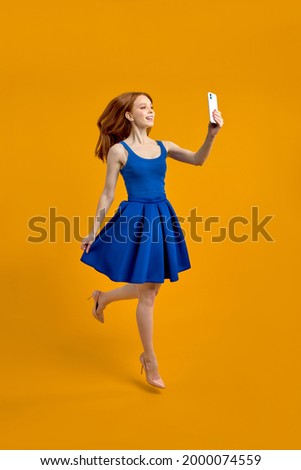 Full length body size photo of stylish trendy redhead woman taking selfie jumping in blue dress isolated yellow studio background, portrait. copy space