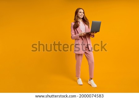 pretty elegant foxy ginger redhaired lady walking carrying laptop isolated on bright yellow orange studio background, copy space. caucasian woman working on laptop, freelance Royalty-Free Stock Photo #2000074583