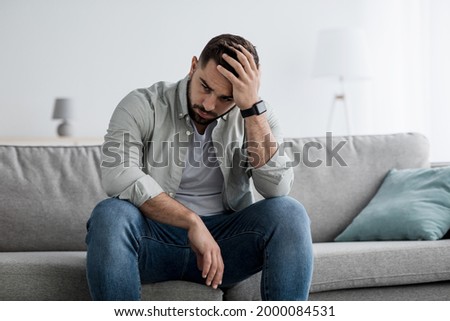 Unhappy man touching hair, sitting on sofa at home, thinking about problems. Upset guy feeling lonely, sad, have psychological and mental troubles, suffering from bad relationship or break up Royalty-Free Stock Photo #2000084531