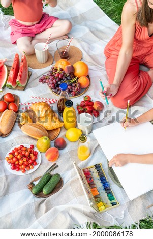 Picnic in summer park, healthy food. Mom with little daughters is engaged in drawing on paper, watercolor paints.