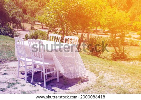 Table covered with a white tablecloth and four white chairs in the garden on the grass on a summer sunny day, toned