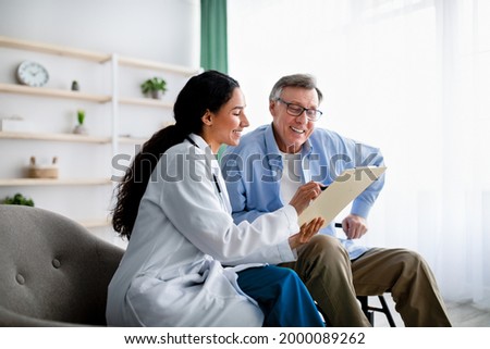 Young doctor asking senior impaired male patient in wheelchair to sign insurance policy at home. Handicapped elderly man putting his signature under surgery consent form, reading medical document Royalty-Free Stock Photo #2000089262