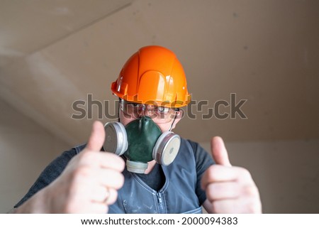 construction worker, a man in an orange helmet and a respirator, shows a thumbs up, a sign of approval, standing against the background of a plasterboard wall at a construction site.