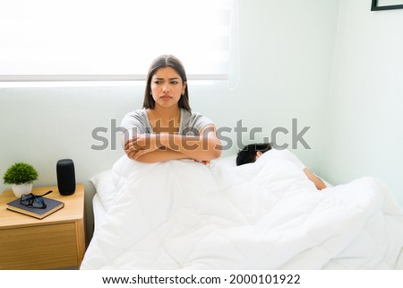 Annoyed hispanic girlfriend sitting up in bed while her boyfriend sleeps next to her. Young woman thinking about the fight with her partner