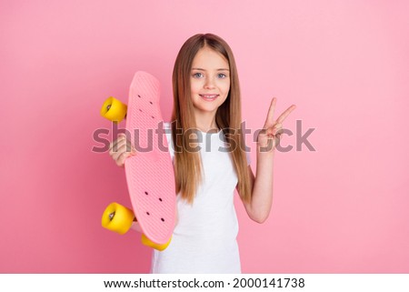 Photo of funny blond little girl show v-sign hold skate wear white t-shirt isolated on pink color background