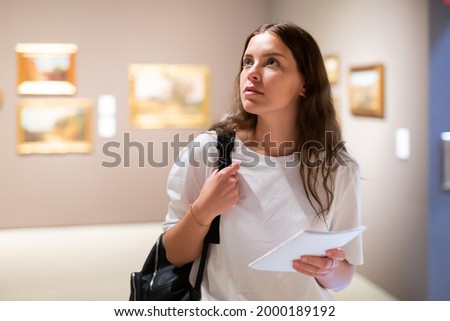 Thoughtful young girl visiting an art gallery exhibition of paintings studies the artwork with information booklet..in her hand Royalty-Free Stock Photo #2000189192