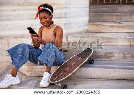 Portrait of happy african-american woman with skateboard. Young stylish woman using the phone outdoors.
