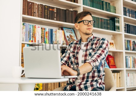 Happy student wearing glasses, portrait of successful man using laptop typing on keyboard, student studying against wall background with books
