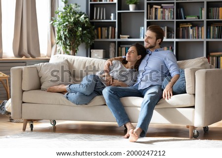 Happy young Caucasian couple renters or tenants relax on couch in living room look in distance dreaming thinking. Smiling dreamy man and woman rest on sofa at home imagine visualize together. Royalty-Free Stock Photo #2000247212