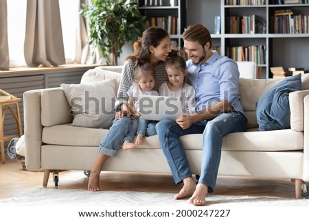 Happy young Caucasian family renters with two small daughters relax in living room with modern laptop. Smiling parents with little girls children rest on sofa at home using computer together.