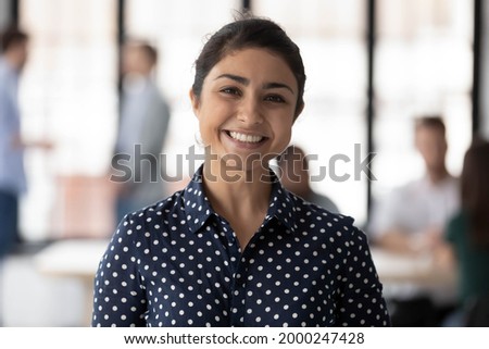 Corporate portrait of happy Indian female business team leader. Millennial employee looking at camera and smiling while group working in office behind her. Businesswoman, leadership concept. Head shot