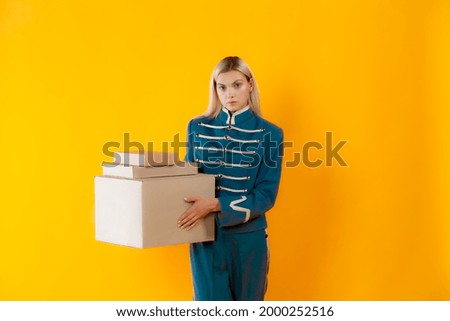 Luxury delivery blond model in hussar coat holding mockup boxes. Photo of slim blonde in old-fashioned military outfit on matte yellow background. Blonde hair girl holding set of mockup boxes.