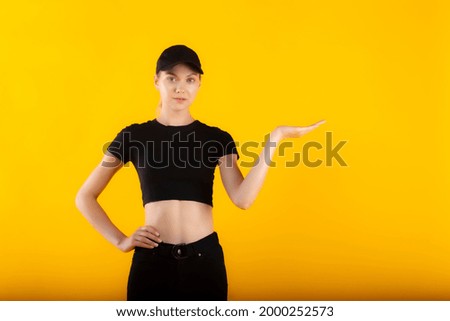 Slim blonde model mockup girl showing of left hand up isolated on yellow. Pretty blond blue-eyed girl model in black high waist jeans, black short t-shirt and black cap posing on yellow matte back