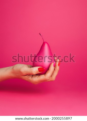 Woman's hand with manicure nails holding a pear painted in pink isolated on a pink background. Creative fruit food photography. Pop art aesthetic.