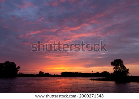Colorful reflection in river IJssel with silhouetted trees of dramatic vibrant intense colorful sunset sky with texture and detail of cloud cover lighting up orange, red and magenta tones. Royalty-Free Stock Photo #2000271548