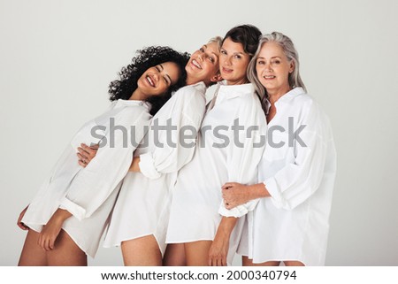 Models of different ages embracing their natural and aging bodies in a studio. Four confident and happy women smiling cheerfully while wearing mens shirts against a white background. Royalty-Free Stock Photo #2000340794
