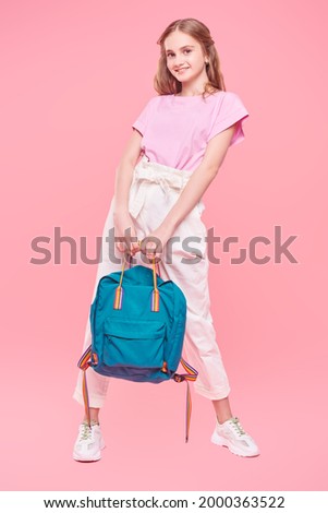 Full length portrait of a carefree pretty girl teenager in light summer clothes and with backpack posing at studio on a pink background. Youth fashion. School time.