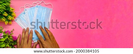 hand holding medical mask on soft pink background with flowers decoration. with the theme of back to school during a pandemic. perfect for cover and banner concepts. copy space, flat lay, top view.