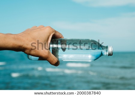 closeup of a young caucasian man holding a glass reusable bottle with water in his hand, in front of the ocean, aligned with the horizon