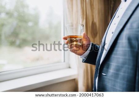 A man in a blue suit, a businessman holds in his hand a glass of whiskey, cognac, rum against the background of the window. Photography, concept.
